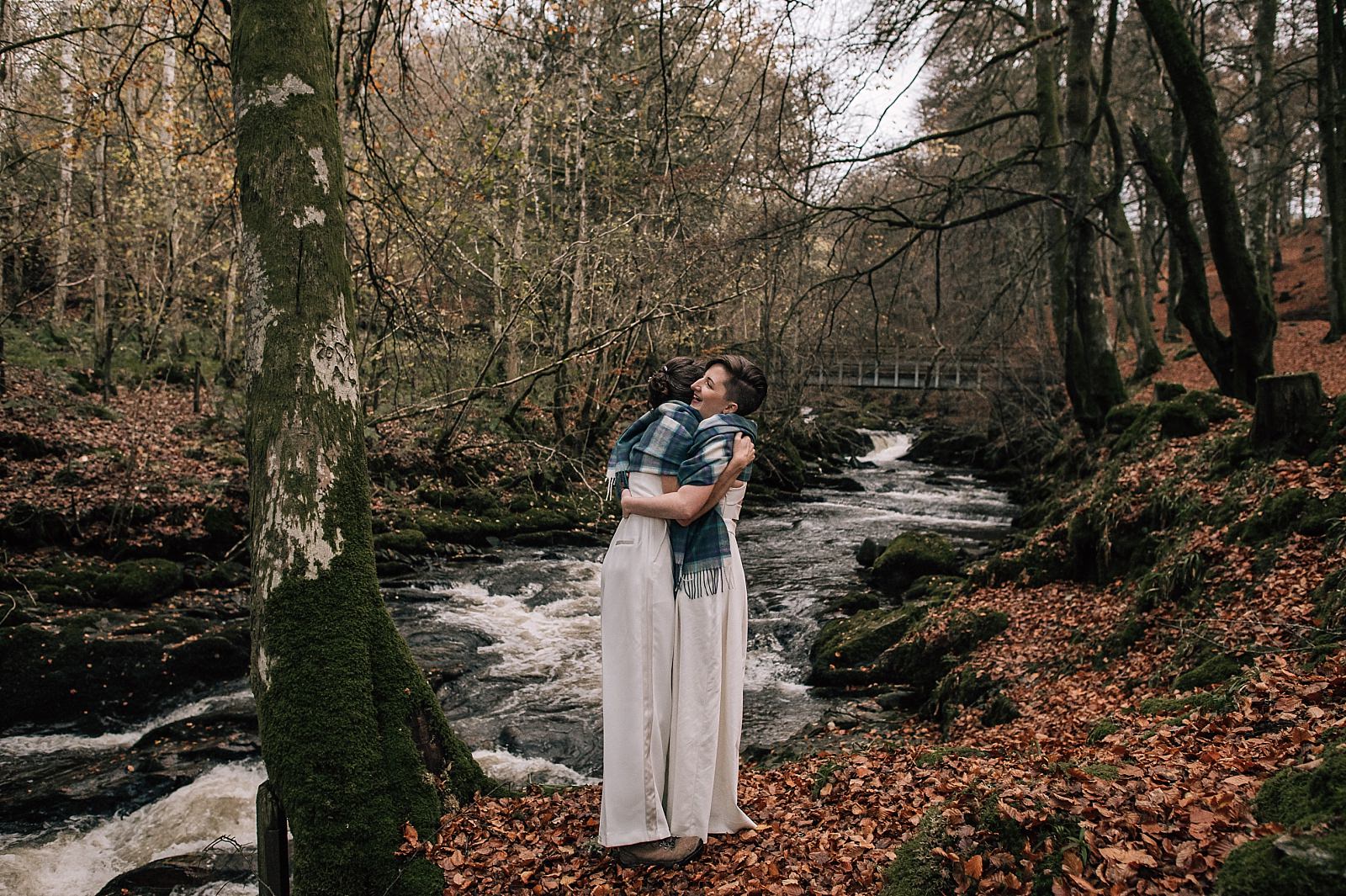 two brides outside in tartan shawls hugging beside a flowing river at boreland loch tay wedding venue aberfeldy perthshire scotland