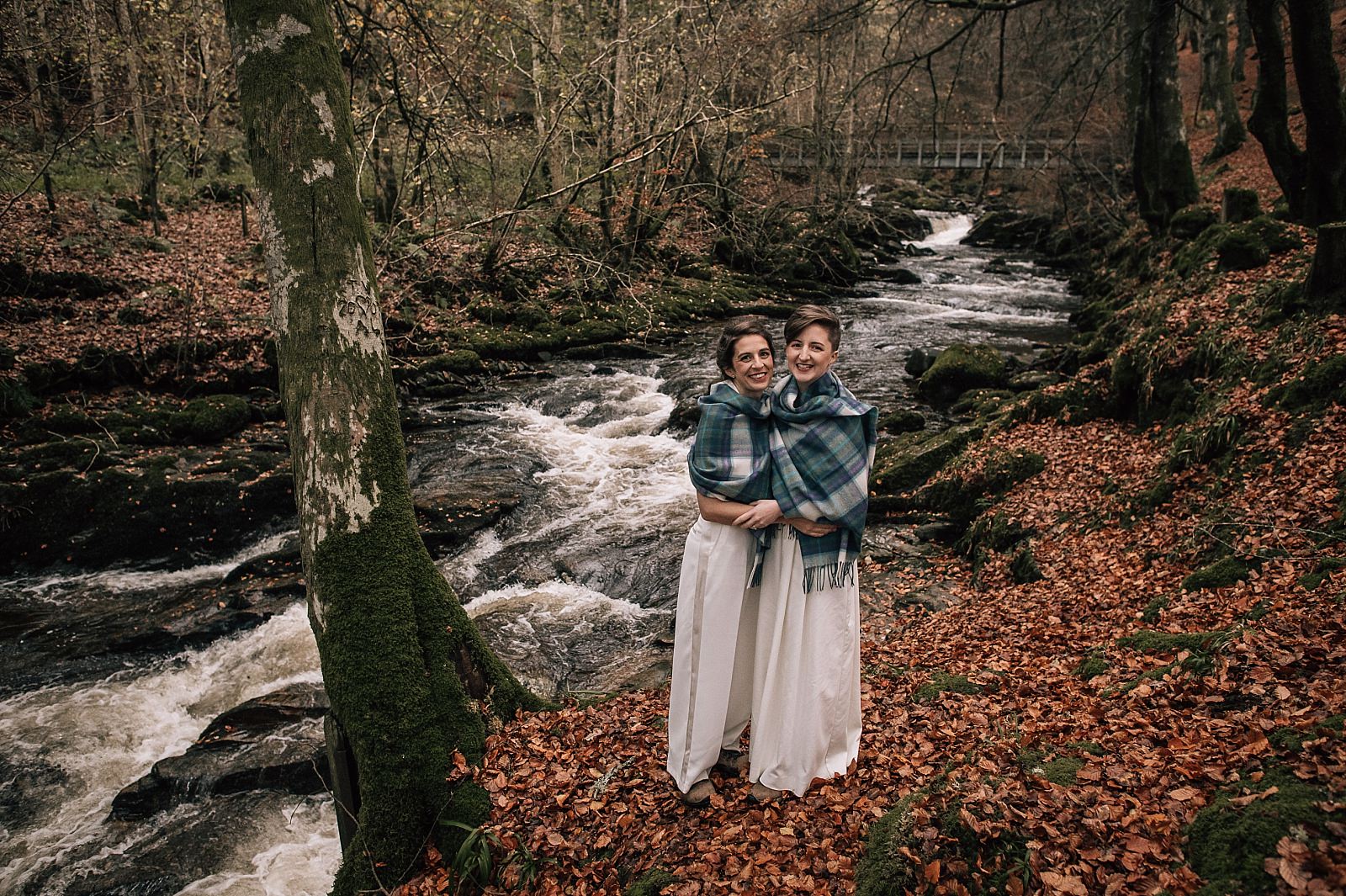 two brides holding each other and smiling beside a flowing river at boreland loch tay wedding venue aberfeldy perthshire scotland