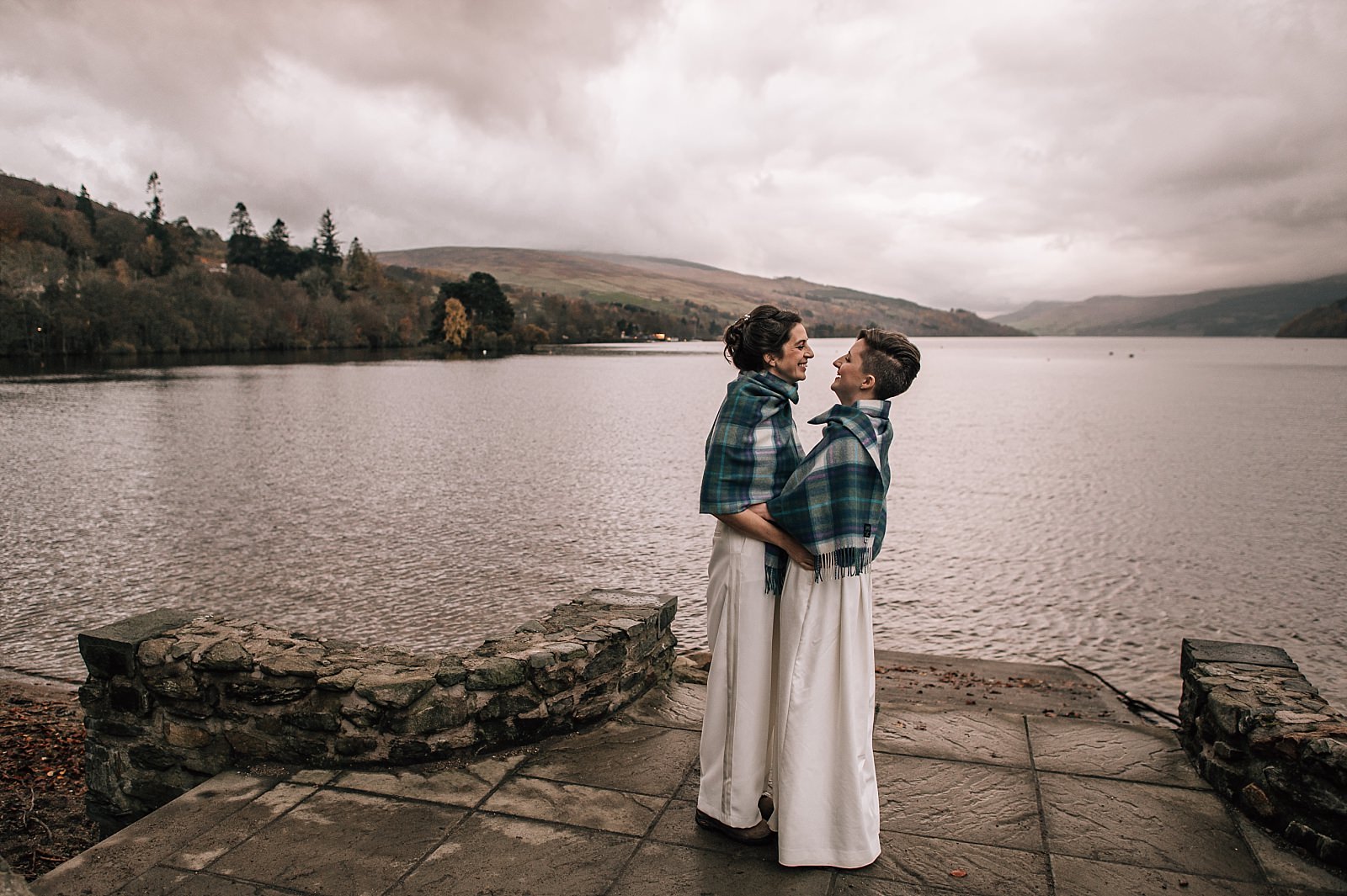 two brides outside in tartan shawls smiling on the shores of boreland loch tay on their wedding day in scotland