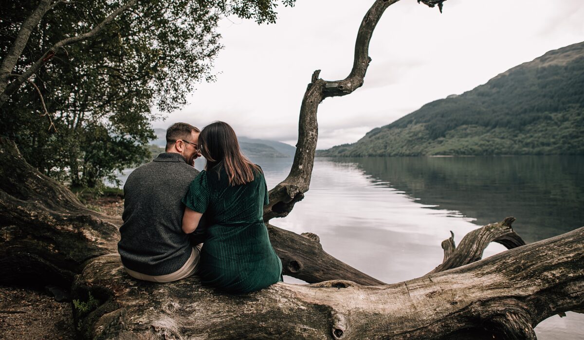 a couple sit side-by-side overlooking scenic lochs and hills at their loch lomond proposal