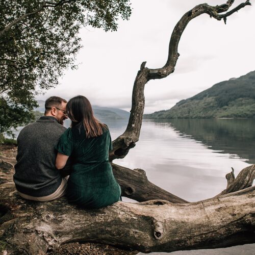 a couple sit side-by-side overlooking scenic lochs and hills at their loch lomond proposal