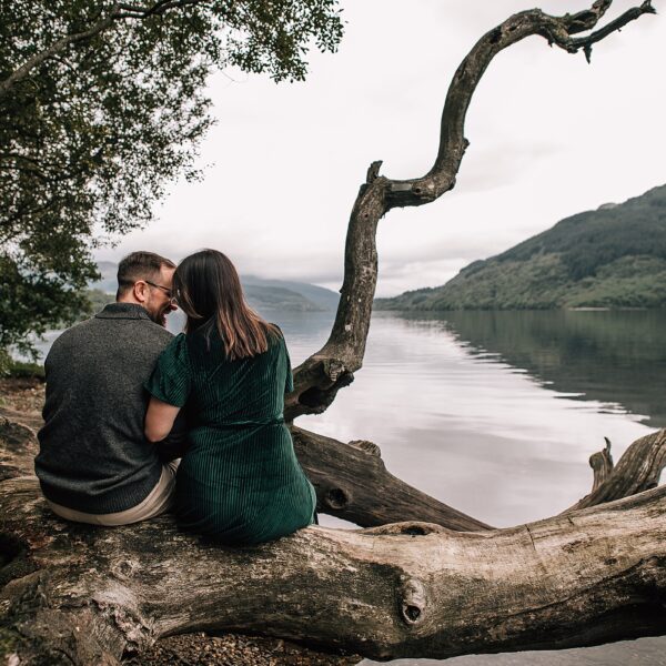 a couple sit side-by-side overlooking scenic lochs and hills at their loch lomond proposal