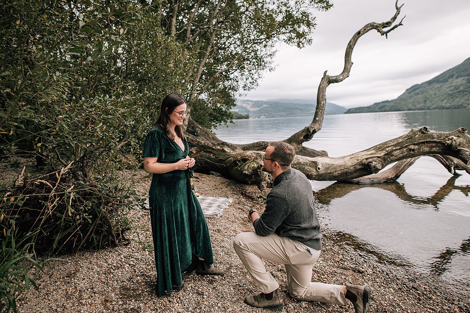 couple standing on shore at Loch Lomond with man down on one knee during a scenic surprise proposal scotland at firkin point