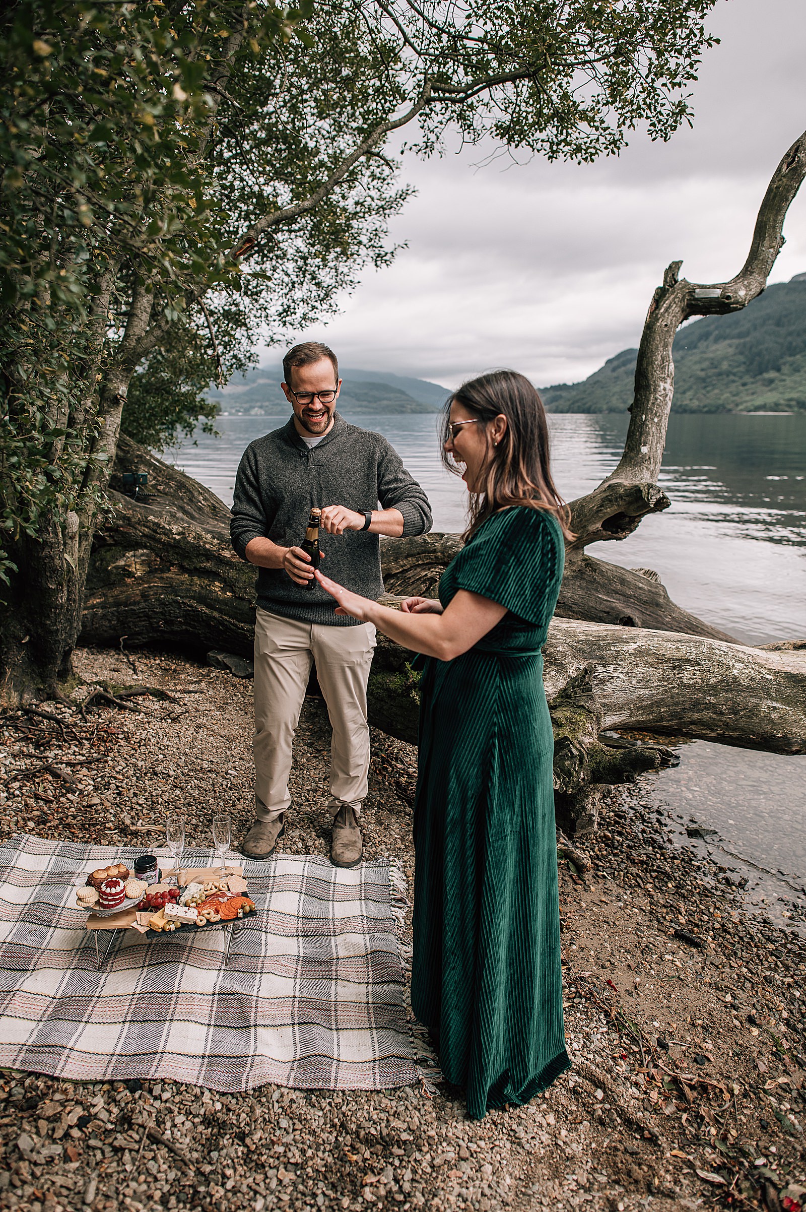 happy and excited newly engaged couple on beach at Loch Lomond firkin point with luxury picnic shot by proposal photographer scotland
