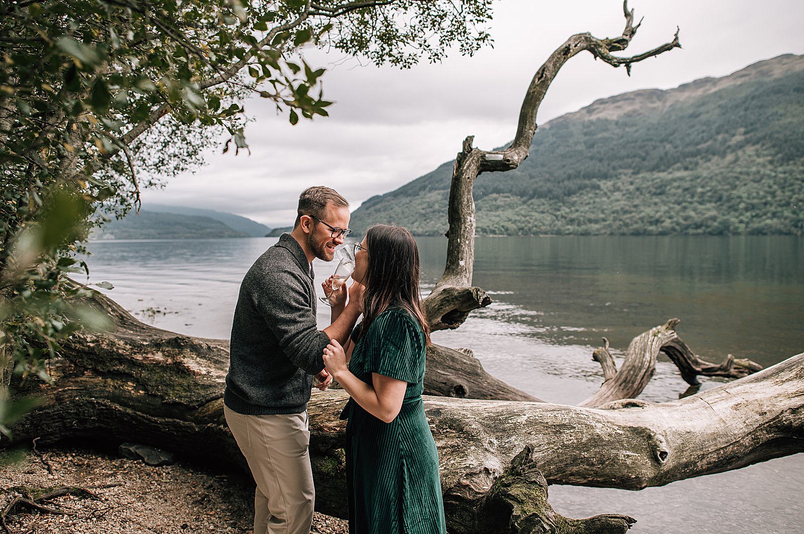 newly engaged couple linking arms and drinking from champagne glasses at their engagement photography scotland shoot with mountains and lochs in scenic scotland
