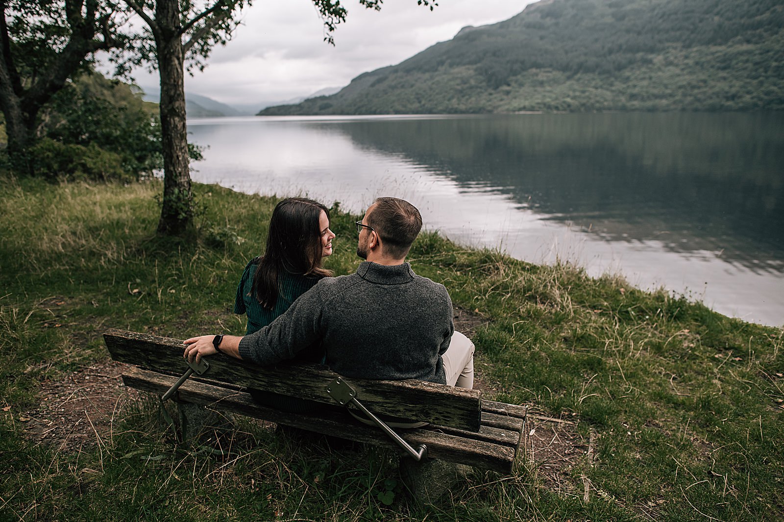 couple sitting on bench at Loch Lomond with very scenic view of the loch and hills after their surprise proposal and engagement photography Scotland photo shoot by proposal photography scotland