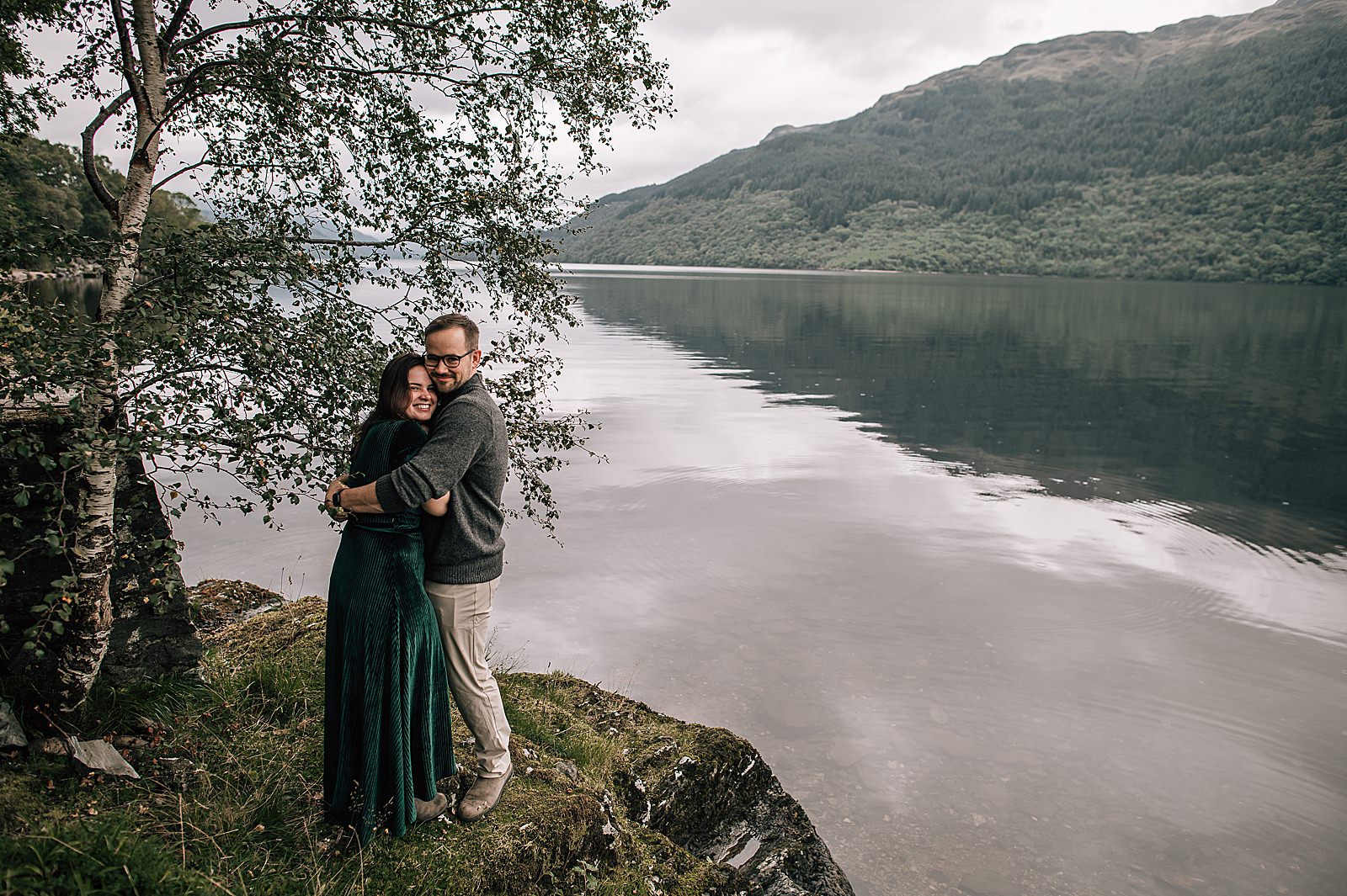 happy smiling couple standing on rock overlooking a tranquil Loch Lomond and a scenic view at their surprise proposal location scotland shot by proposal photographer scotland