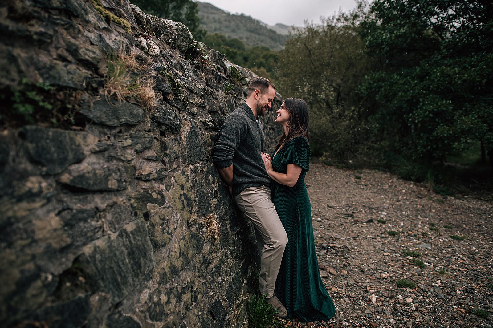 happy couple standing against stone wall at Loch Lomond after their surprise proposal shot by proposal photographer scotland
