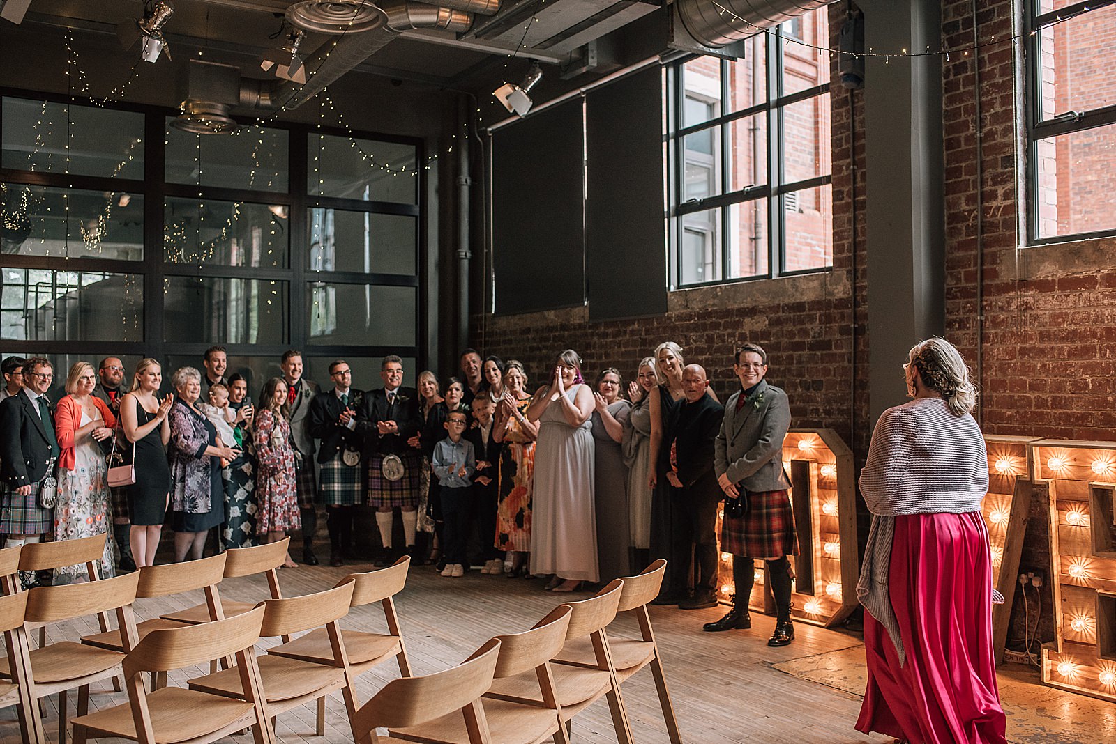 bride walking in pink wedding dress for first look in front of guests at west brewery wedding with fairy lights wooden chairs and large light up love sign