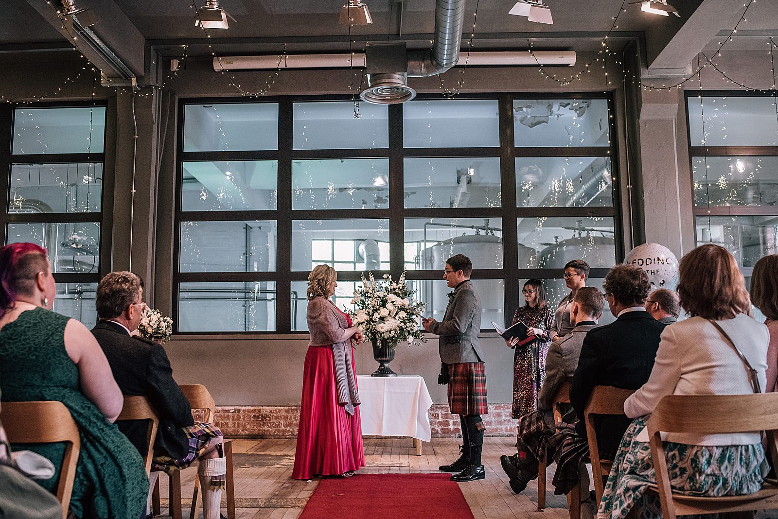 bride and groom during their vows at west brewery wedding standing in front of large window with view of west beer brewery and fairy lights hanging above industrial space for wedding glasgow