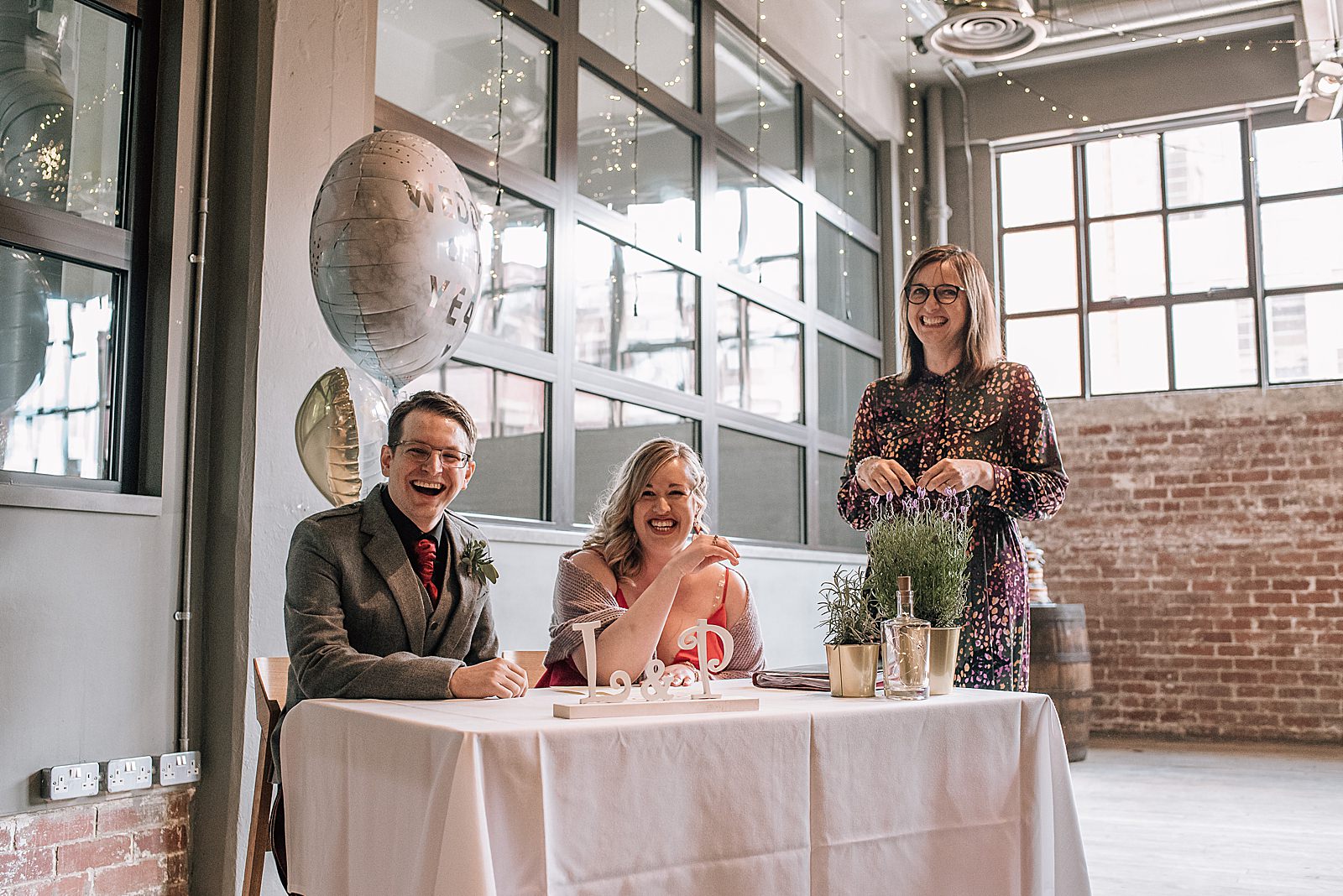 bride and groom sitting at table to sign marriage certificate at west brewery wedding looking at the camera and smiling with celebrant inside west brewery