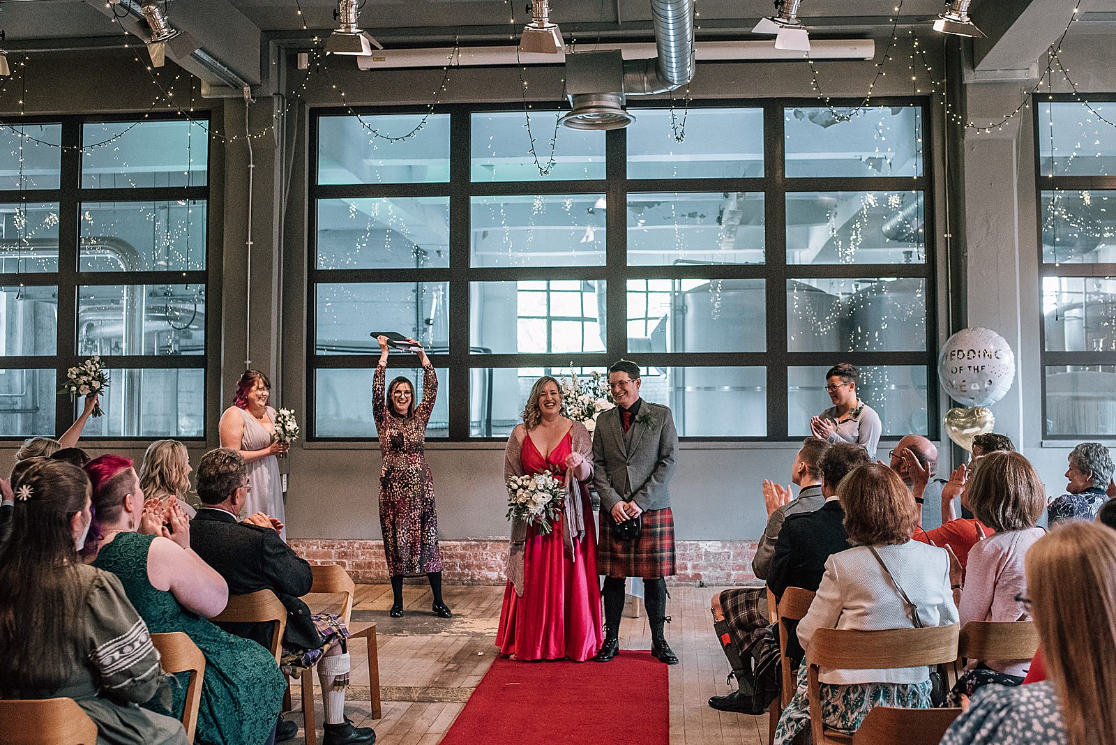 bride and groom smiling while celebrant cheers in front of guests at the end of their ceremony during west brewery wedding with groom dressed in a kilt and bride in a satin pink wedding dress for informal wedding shot by alternative wedding photographer glasgow