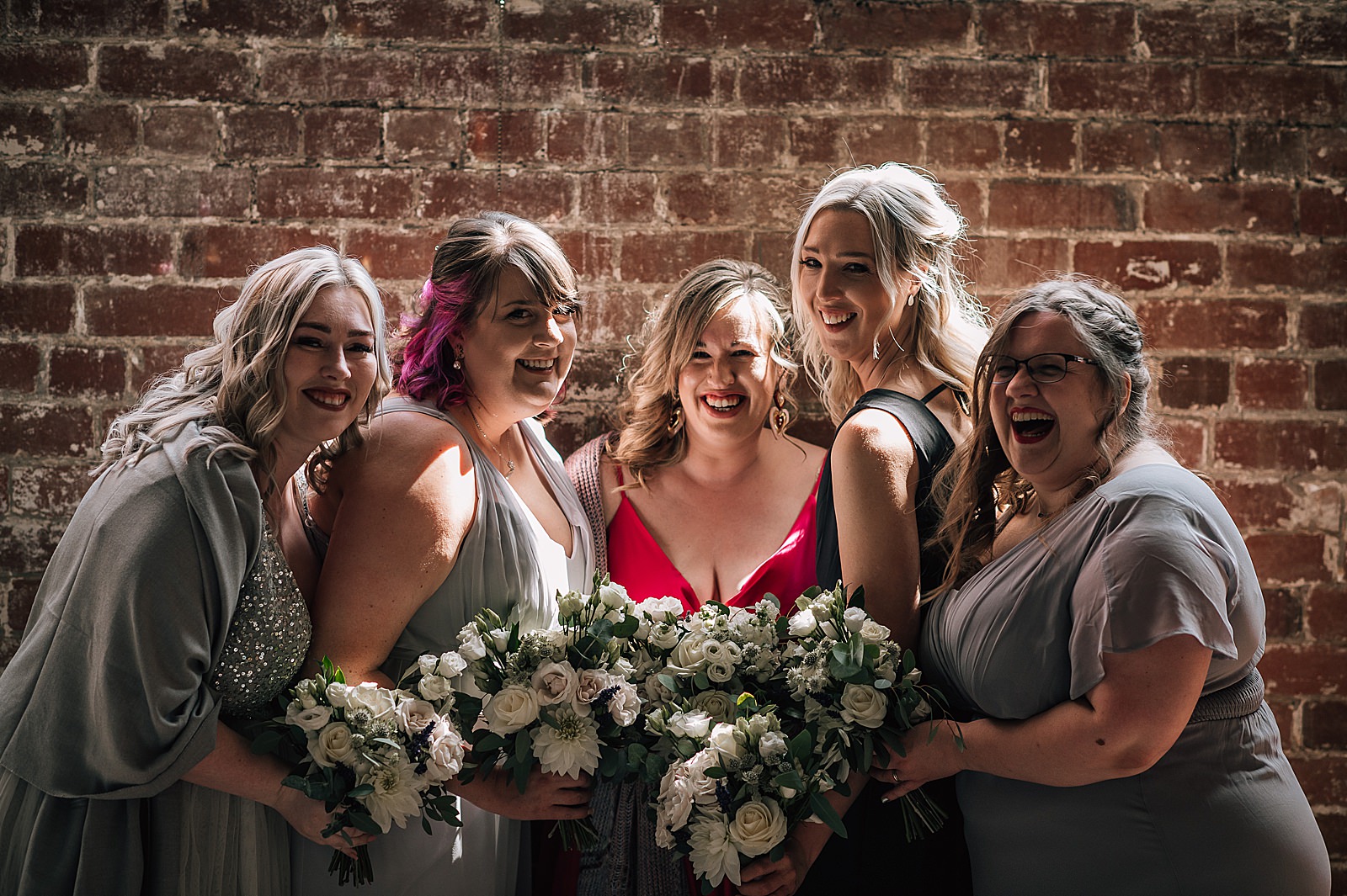 bride and her bridesmaids holding white flower bouquets smiling while standing against brick wall with beautiful natural light after west brewery wedding ceremony