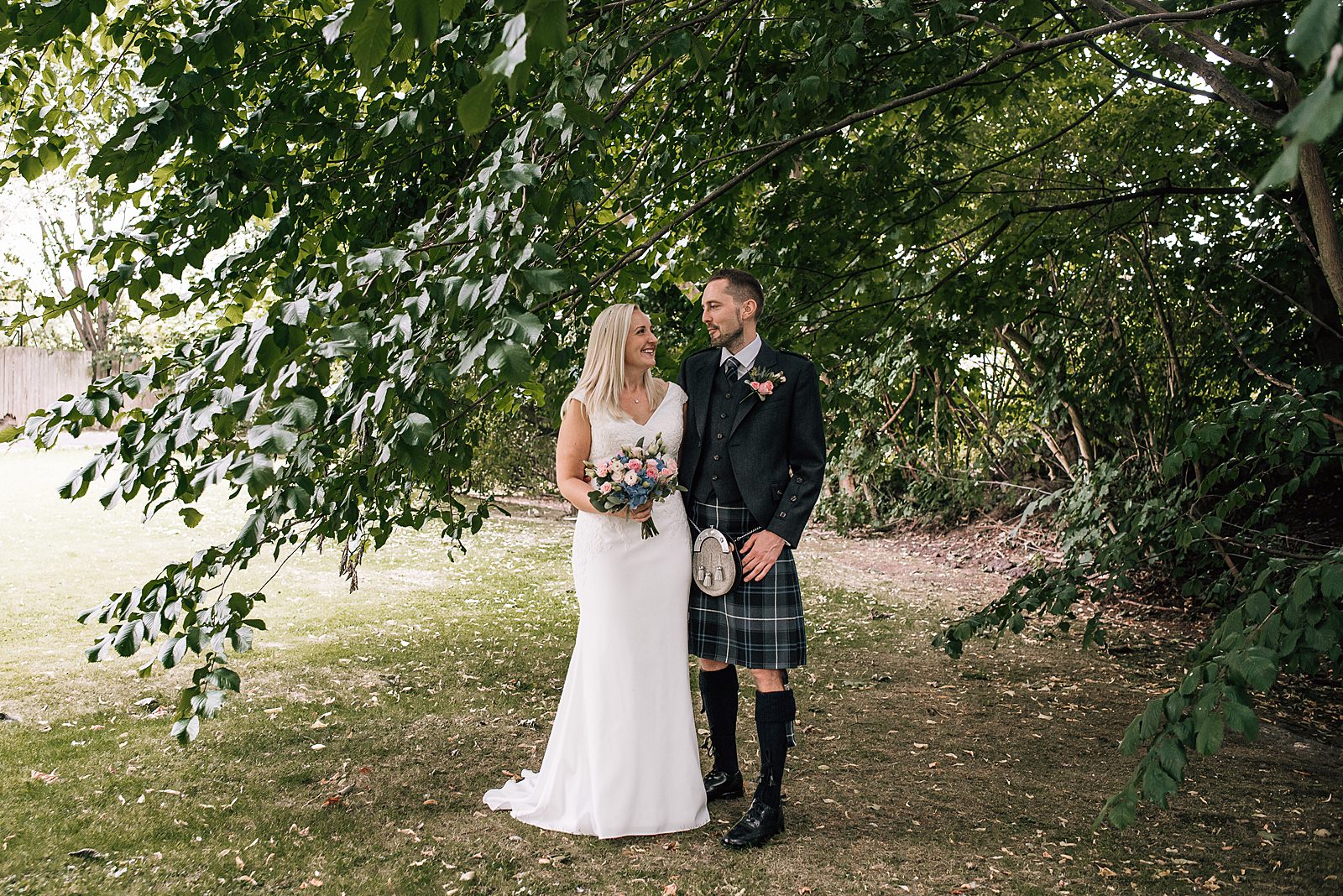 bride and groom in garden under lush green tree looking at each other and smiling on morning of their the bothy glasgow wedding