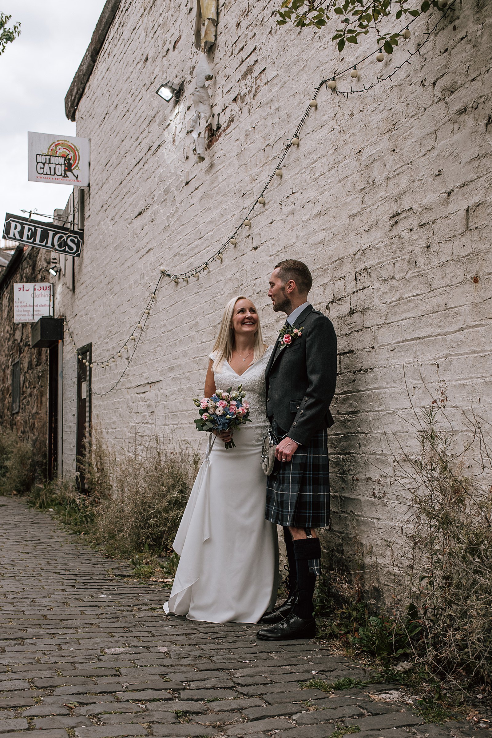 bride and groom standing against white brick wall looking at each other and smiling near the bothy glasgow after west end wedding