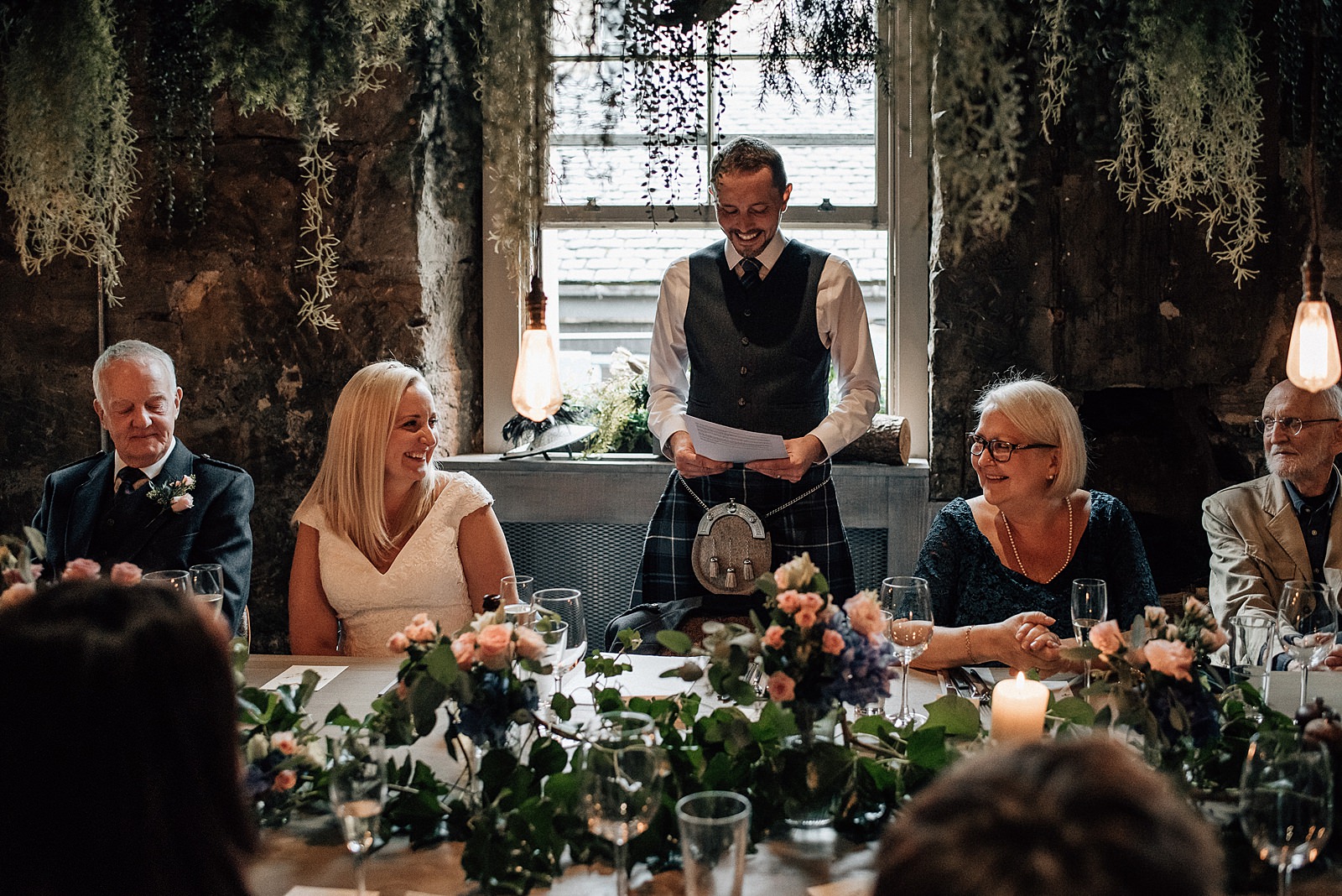 groom standing in front of window making speech at the bothy Glasgow wedding as bride laughs looking at her mother