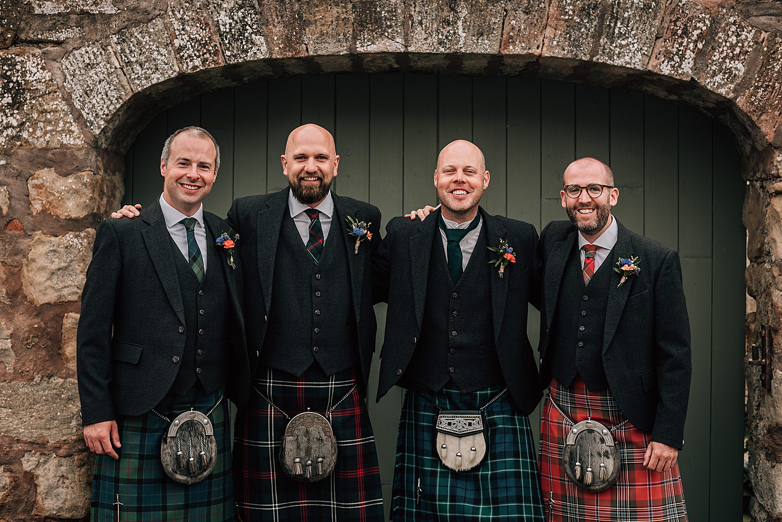 groomsmen standing together and smiling underneath arched doorway at the cowshed Scotland in crail