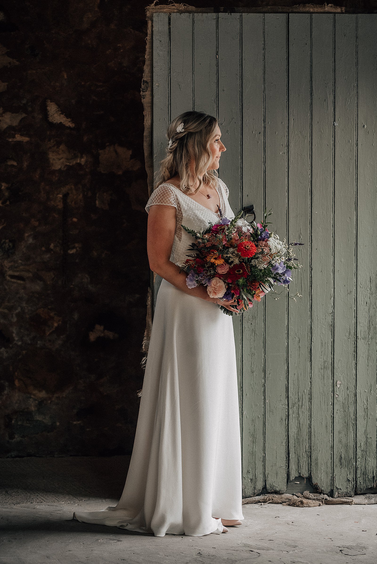 bridal portrait of bride standing in flowy white dress holding a colourful wild flower bouquet inside barn at the cow shed scotland in crail fife