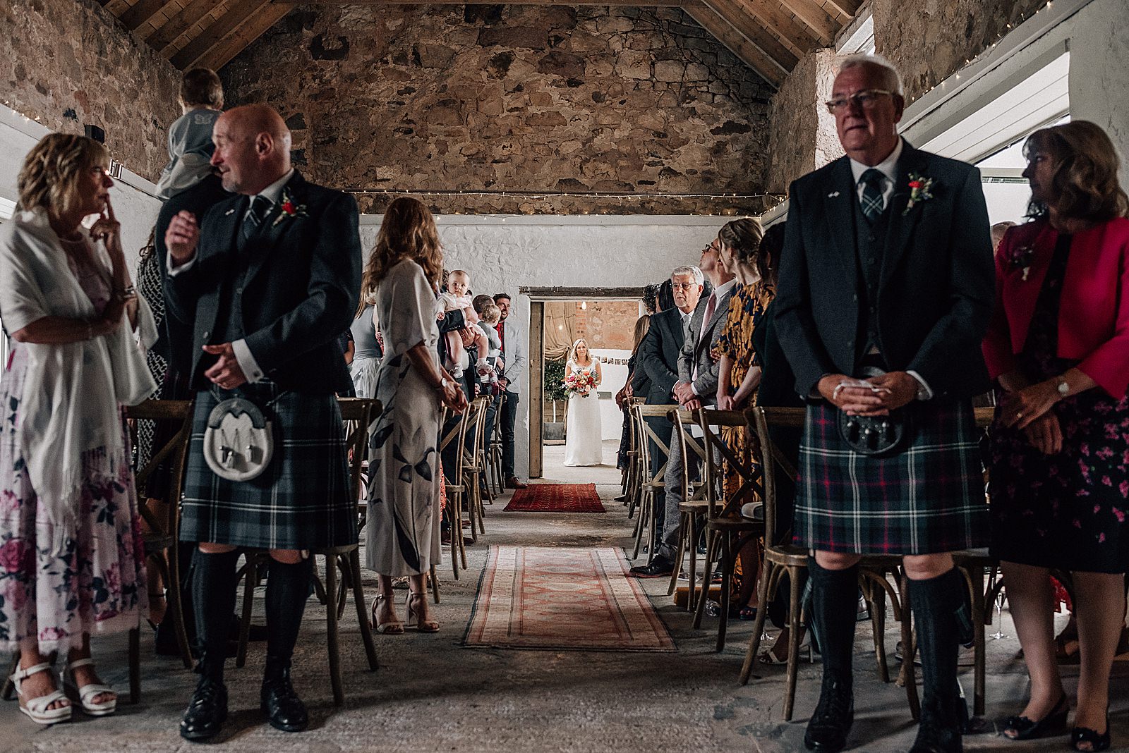bride standing in doorway waiting to walk down the aisle as wedding guests are standing and waiting for her inside rustic barn at the cow shed scotland