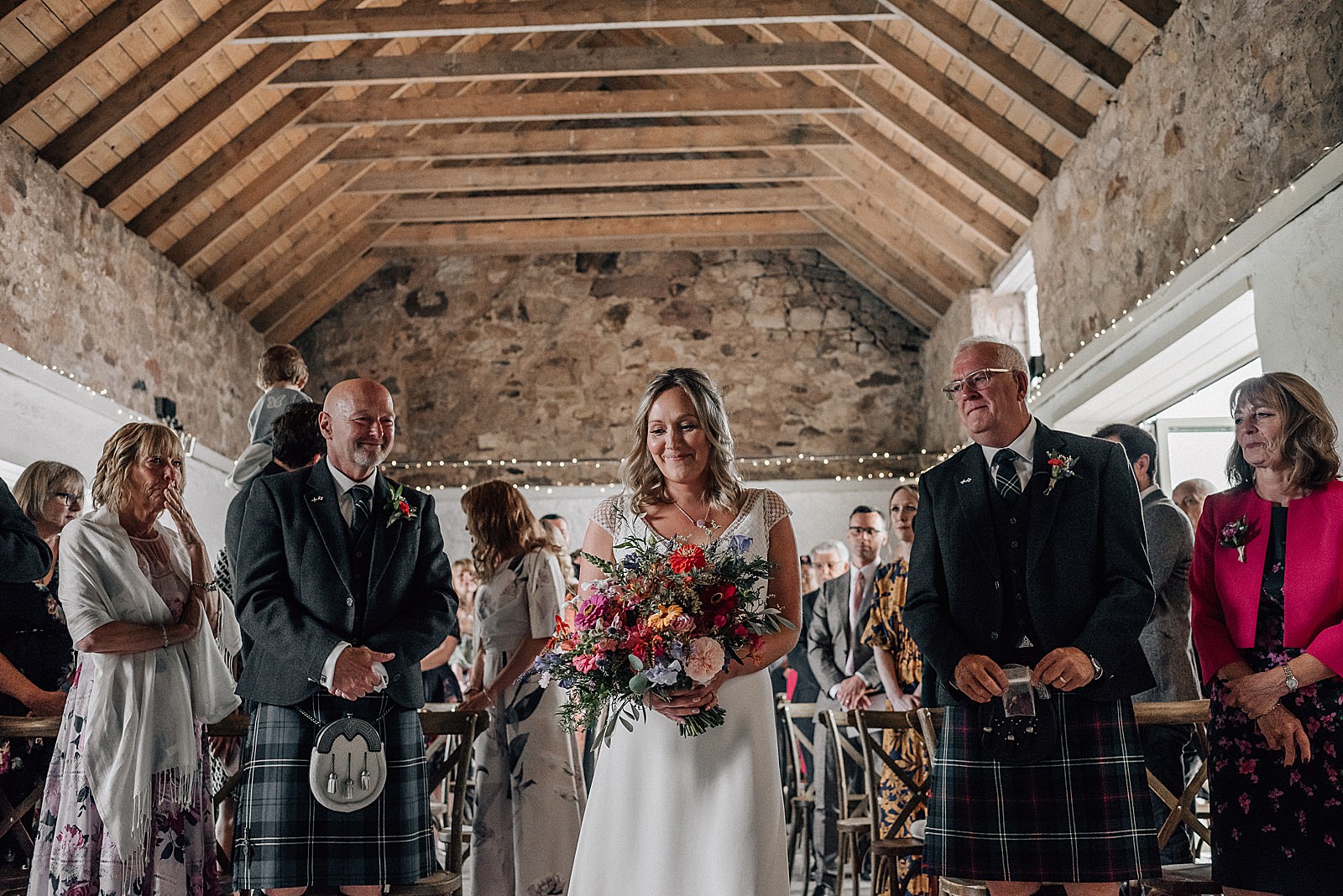 bride walking herself down the aisle holding a colourful wild flower bouquet inside rustic barn for her diy Scottish wedding at the cow shed scotland