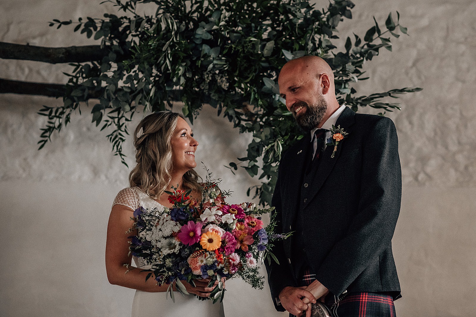 bride and groom smiling at each other standing in front of green foliage arch and rustic white barn wall during their diy Scottish wedding