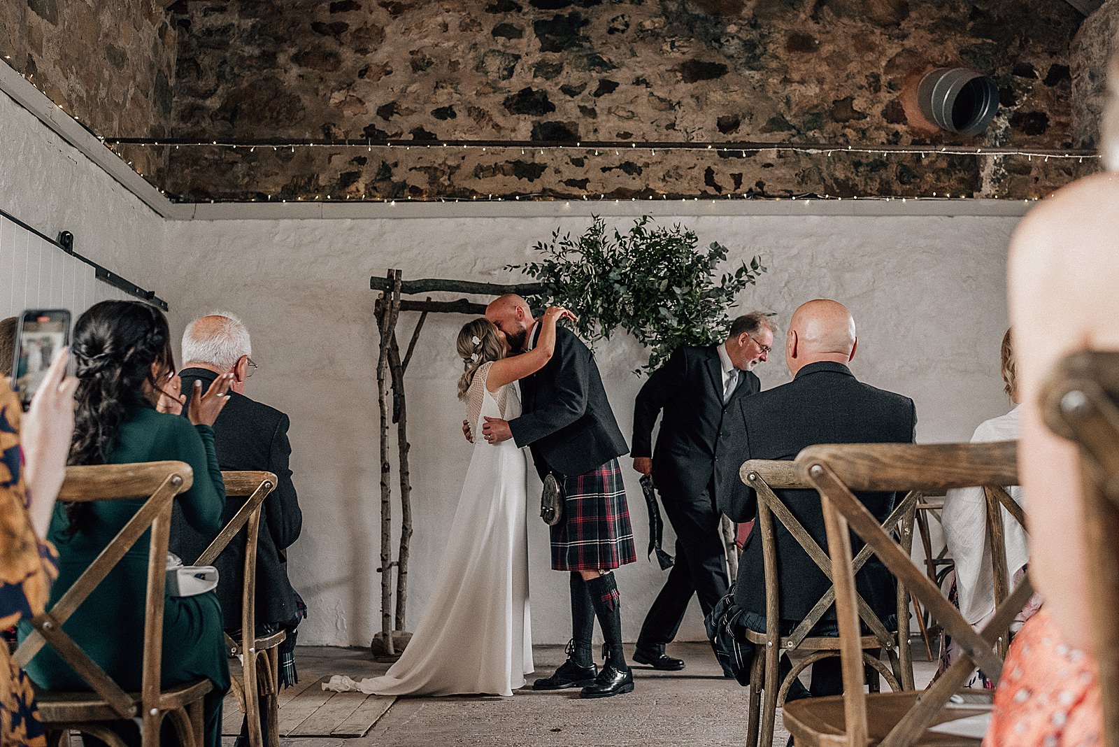bride and groom's first kiss under green foliage arch inside barn with rustic walls for their wedding at cow shed crail in St Andrews