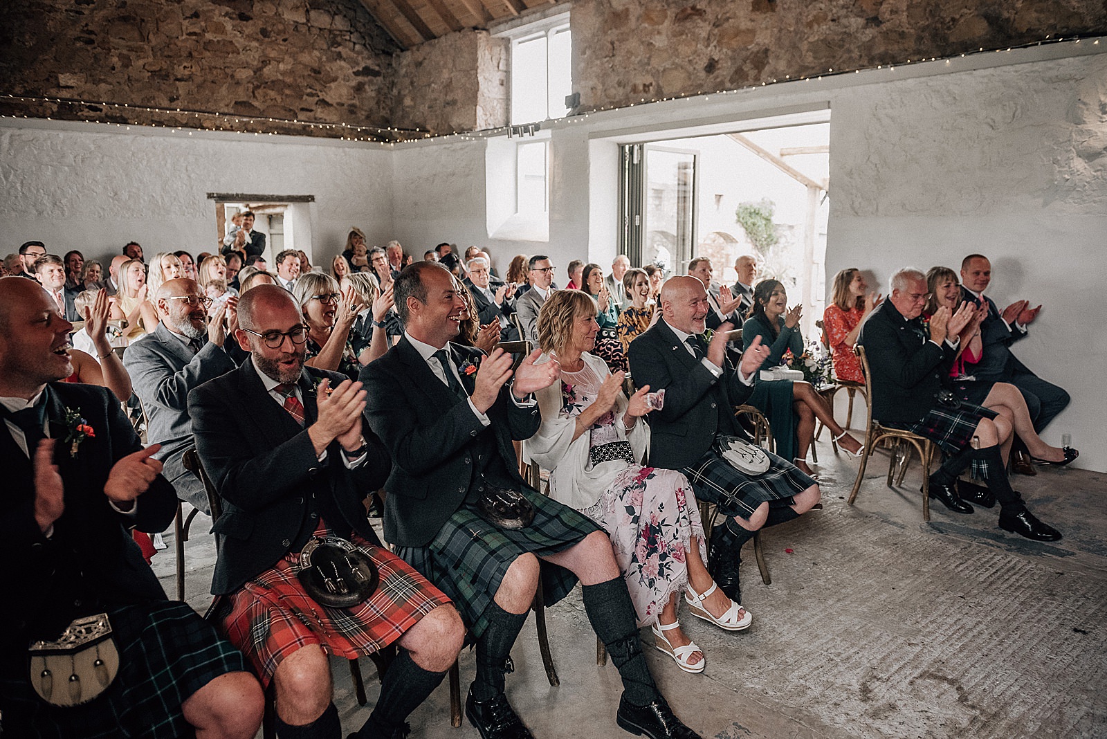 happy wedding guests cheering and clapping at the end of wedding ceremony at the cow shed scotland inside rustic barn wedding scotland