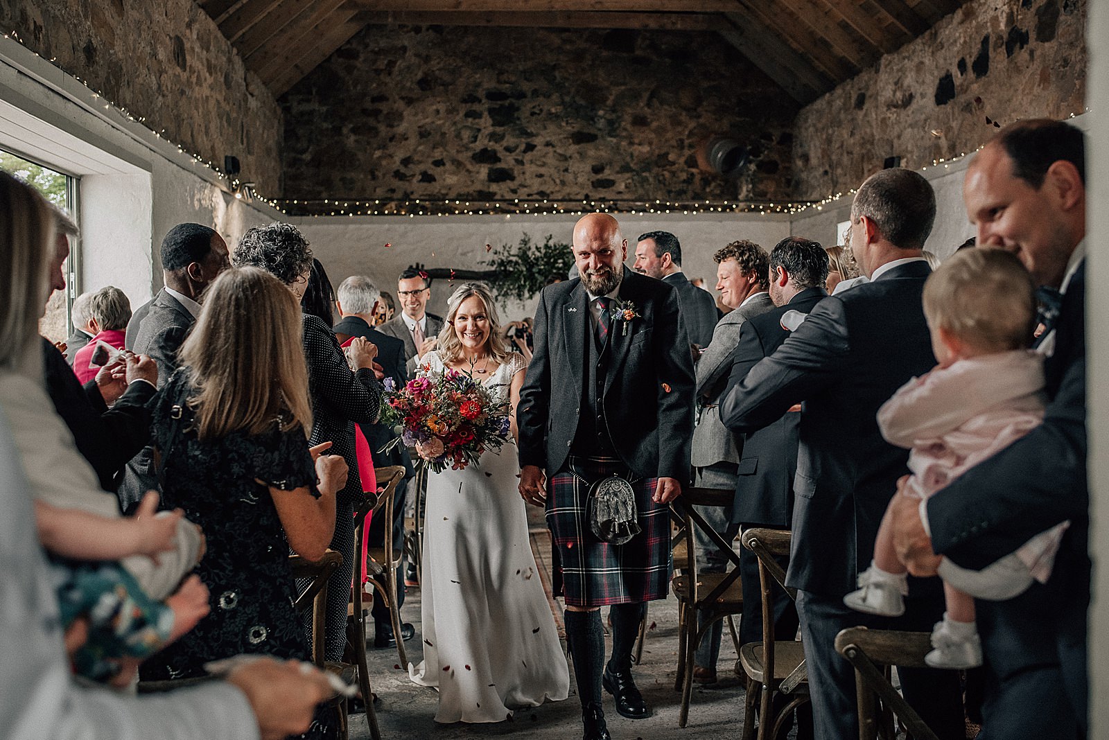 bride and groom smiling and walking under confetti throw inside rustic barn at the cow shed scotland after their quirky wedding