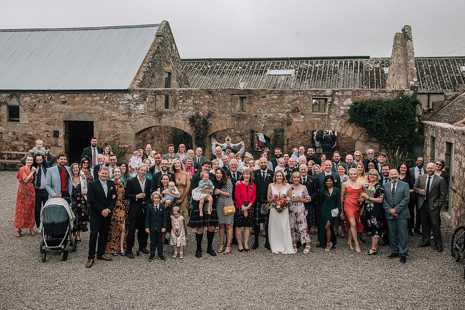 happy wedding guests gathered for big group shot in courtyard at the cow shed scotland, a wedding venue in St Andrews