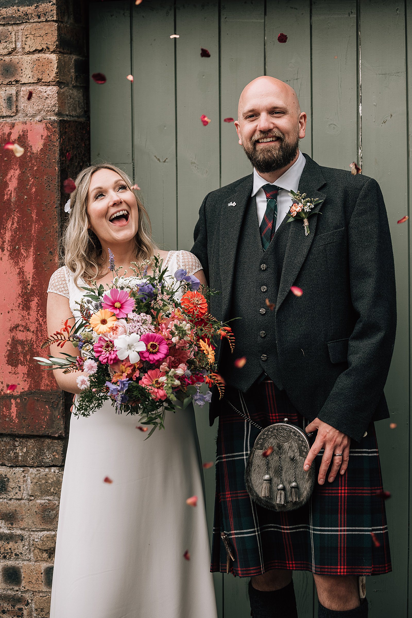 bride and groom happy and smiling standing in front of green barn door under a rose petal confetti throw at the cow shed scotland