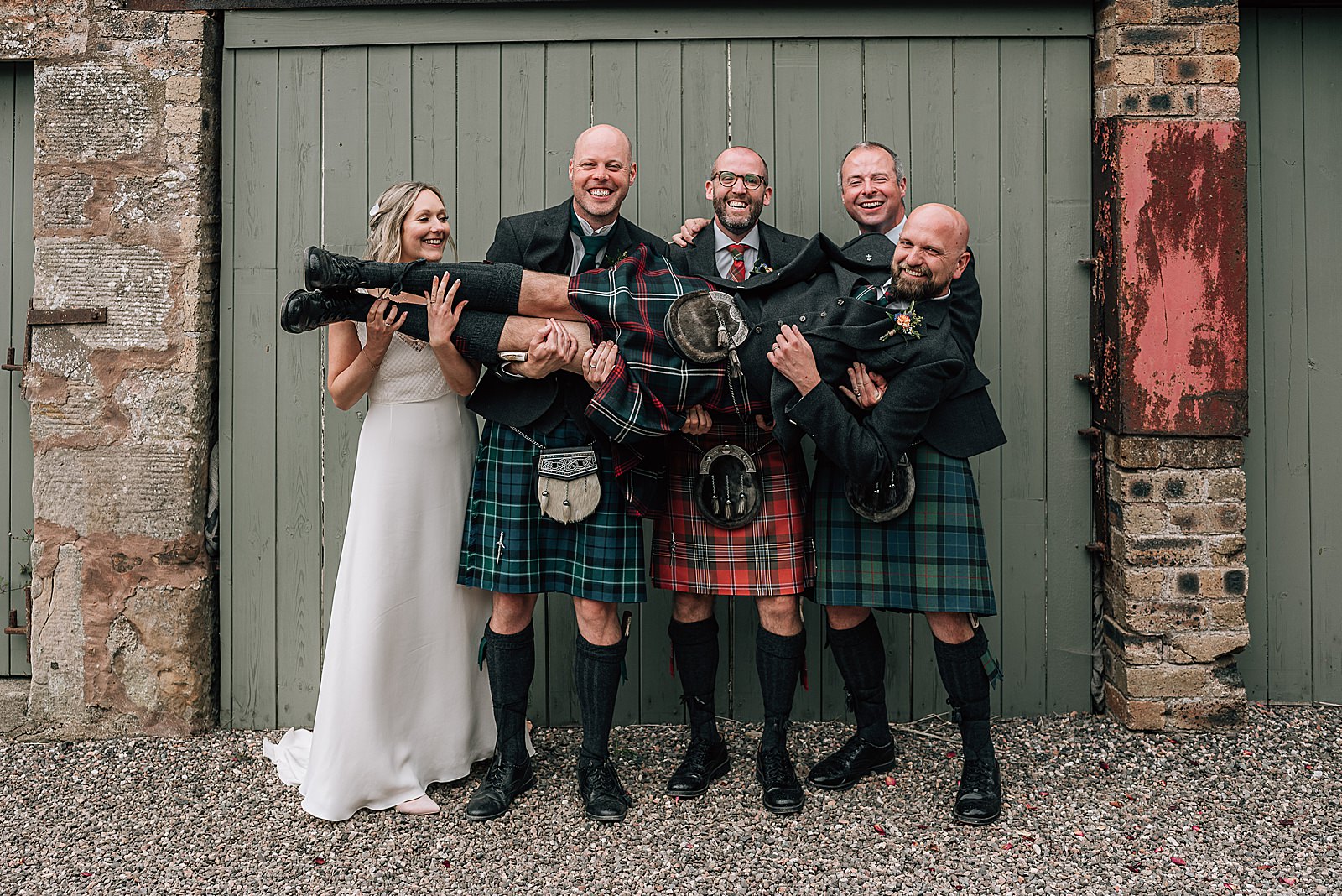 groomsmen and bride all holding groom dressed in kilt while smiling and laughing in front of green barn door at the cow shed crail after their quirky wedding scotland