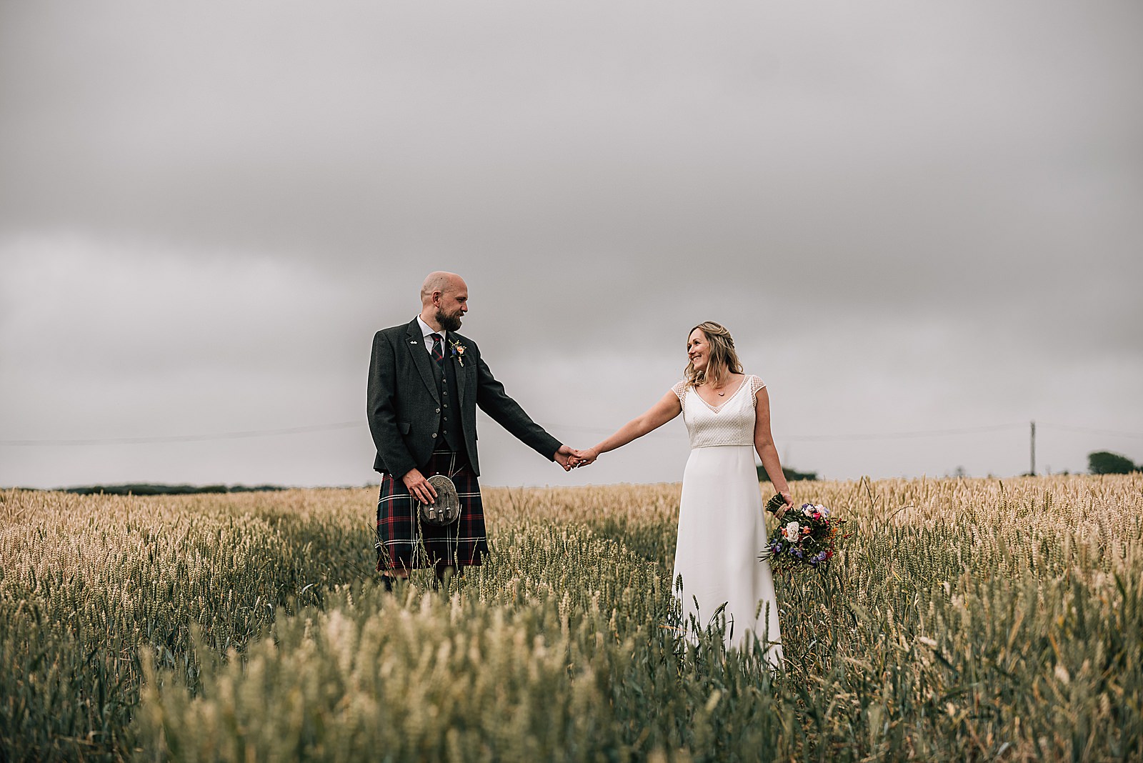 bride and groom walking through wheat field and smiling at each other during their diy Scottish wedding at the cow shed crail