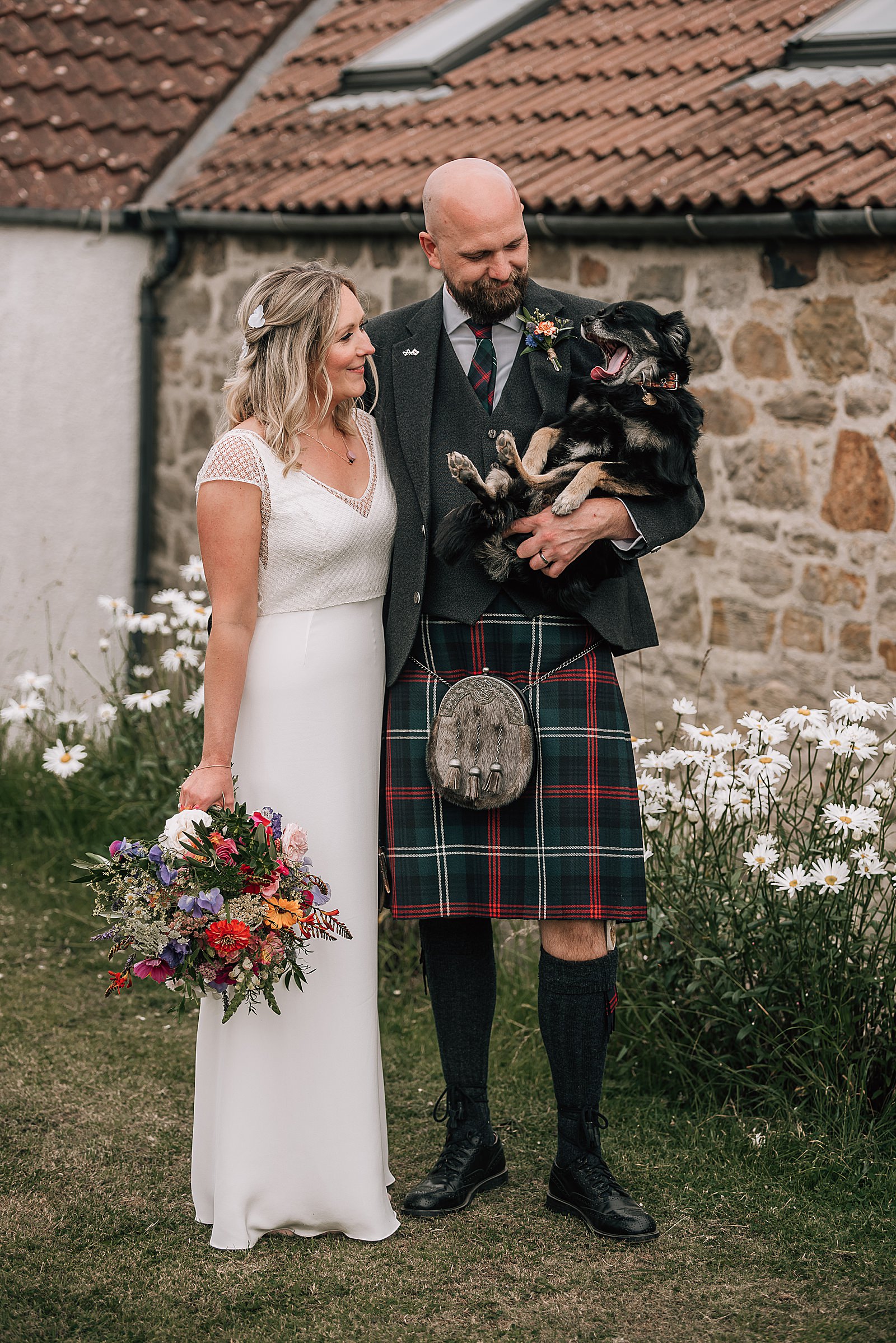 bride and groom standing and cuddling their dog outside rustic barn at the cow shed scotland after their diy Scottish wedding