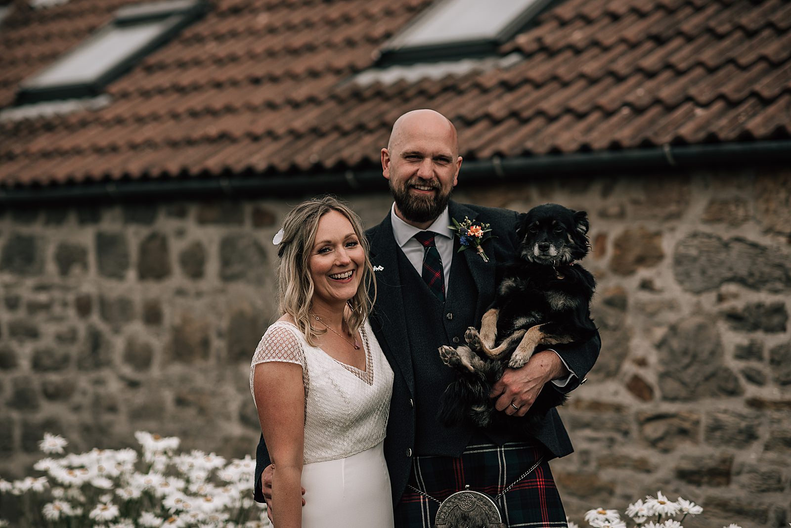 bride and groom smiling and cuddling their dog outside rustic barn at the cow shed scotland after their diy Scottish wedding