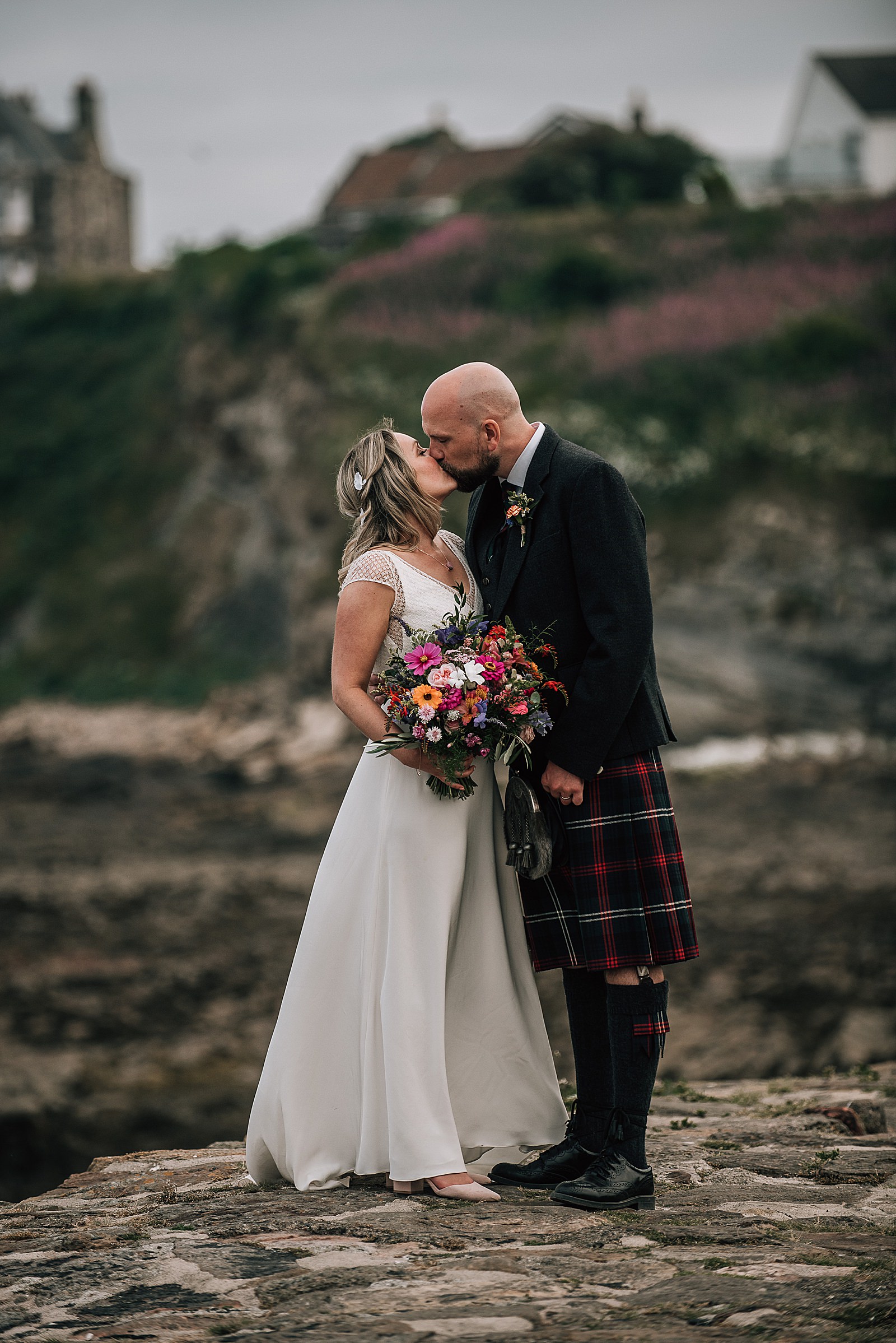 bride in a flowy boho white dress and groom in highland dress in kilt at crail harbour kissing after their wedding at the cow shed scotland