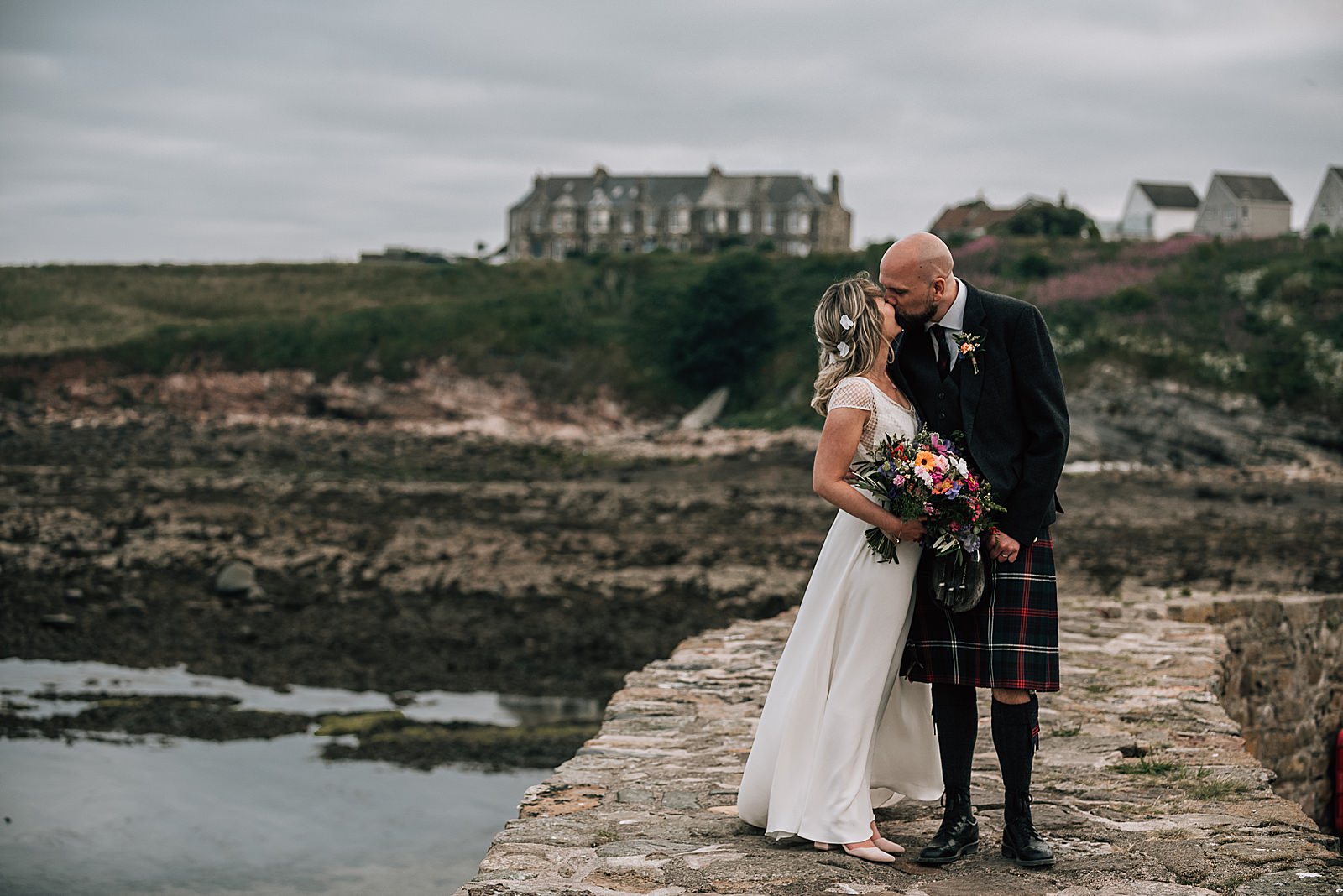 bride in a flowy boho white dress and groom in highland dress in kilt at crail harbour kissing after their quirky Scottish wedding at the cow shed scotland
