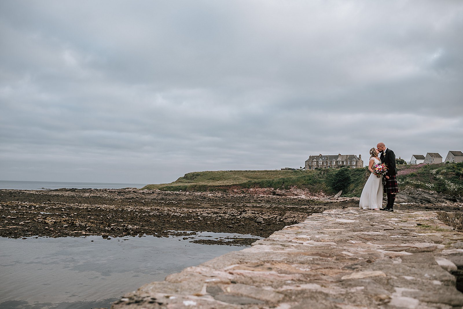 bride in a flowy boho white dress and groom in kilt at crail harbour kissing after their wedding at the cow shed