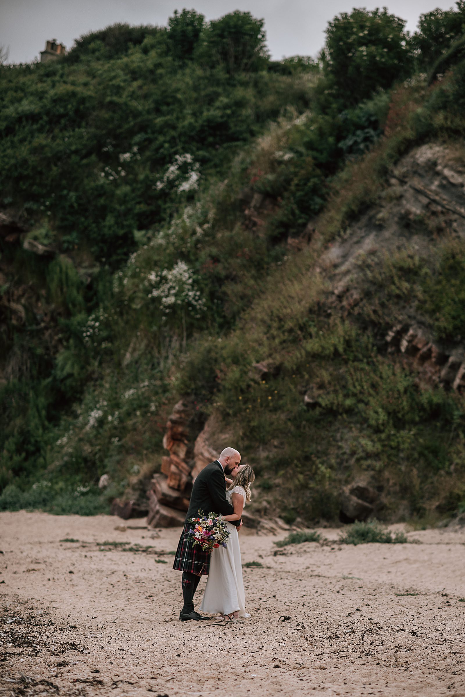 bride and groom kissing standing on the beach near the cow shed scotland underneath beautiful green cliff