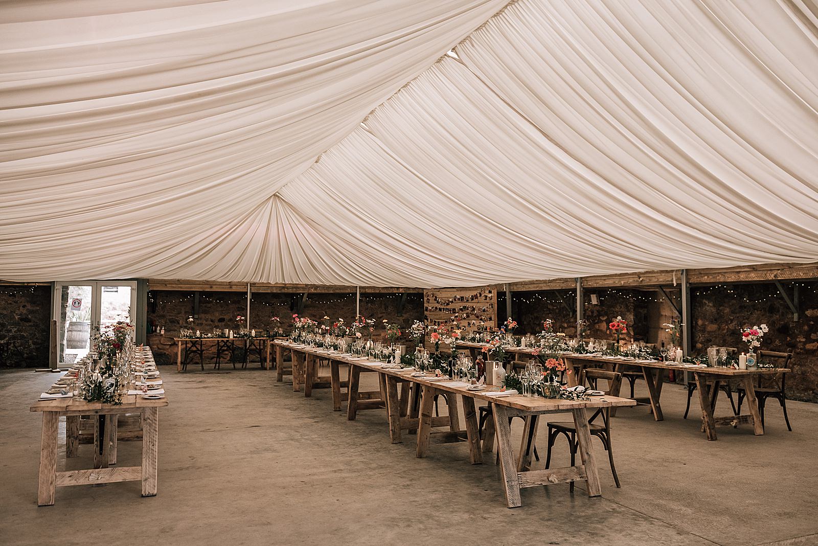 reception set up for wedding at the cow shed crail with rustic wooden tables and flowers underneath tent roof with fairy lights during a diy Scottish wedding