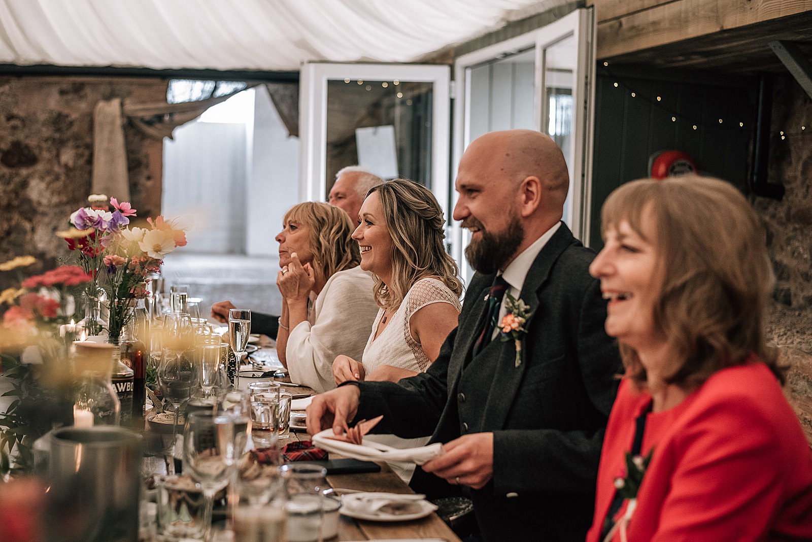 bride and groom with family smiling and laughing at top table during speeches at the cow shed scotland for their diy Scottish wedding