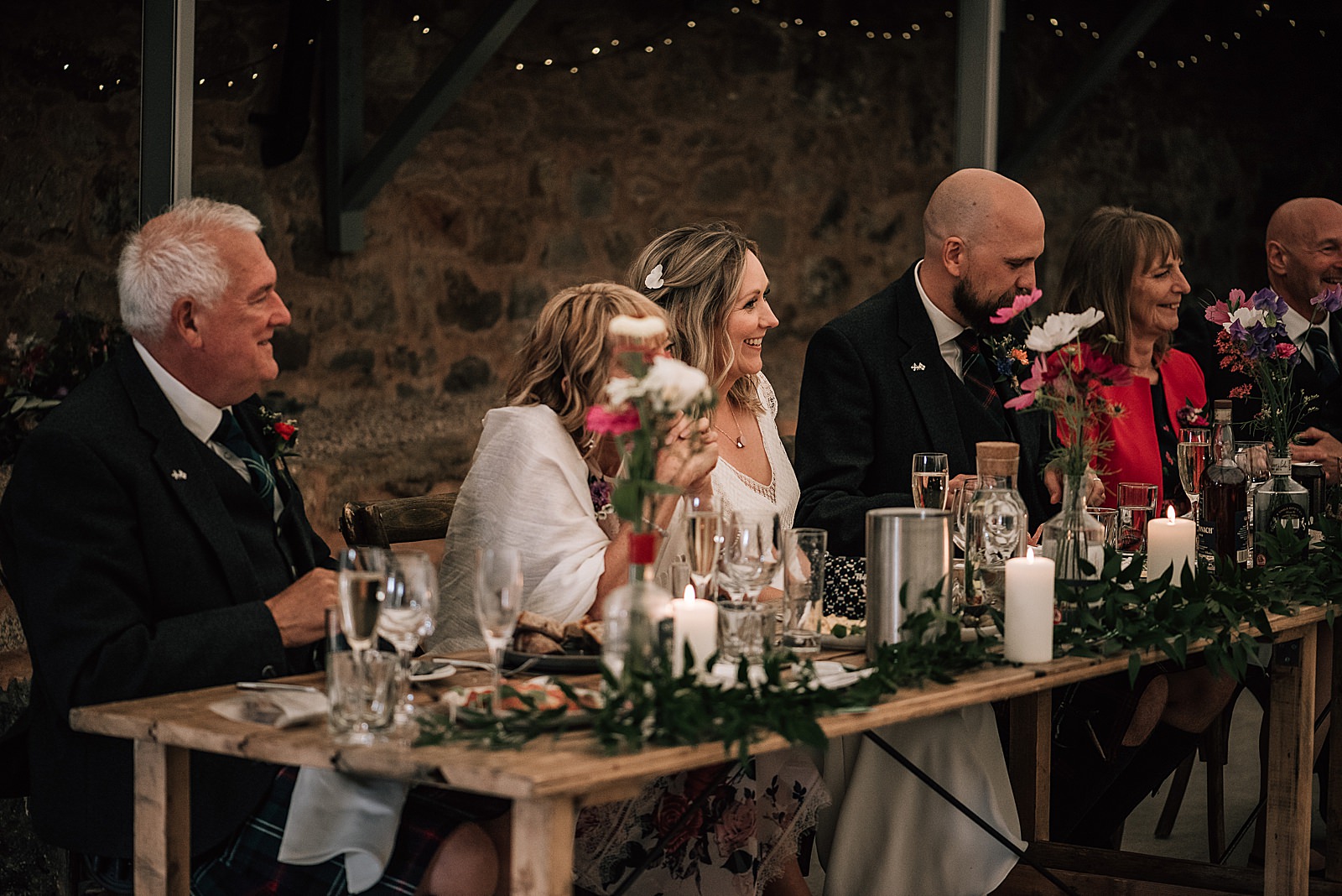 bride and groom with family smiling and laughing at top table during speeches at the cow shed scotland for their diy Scottish wedding with beautiful pink flowers