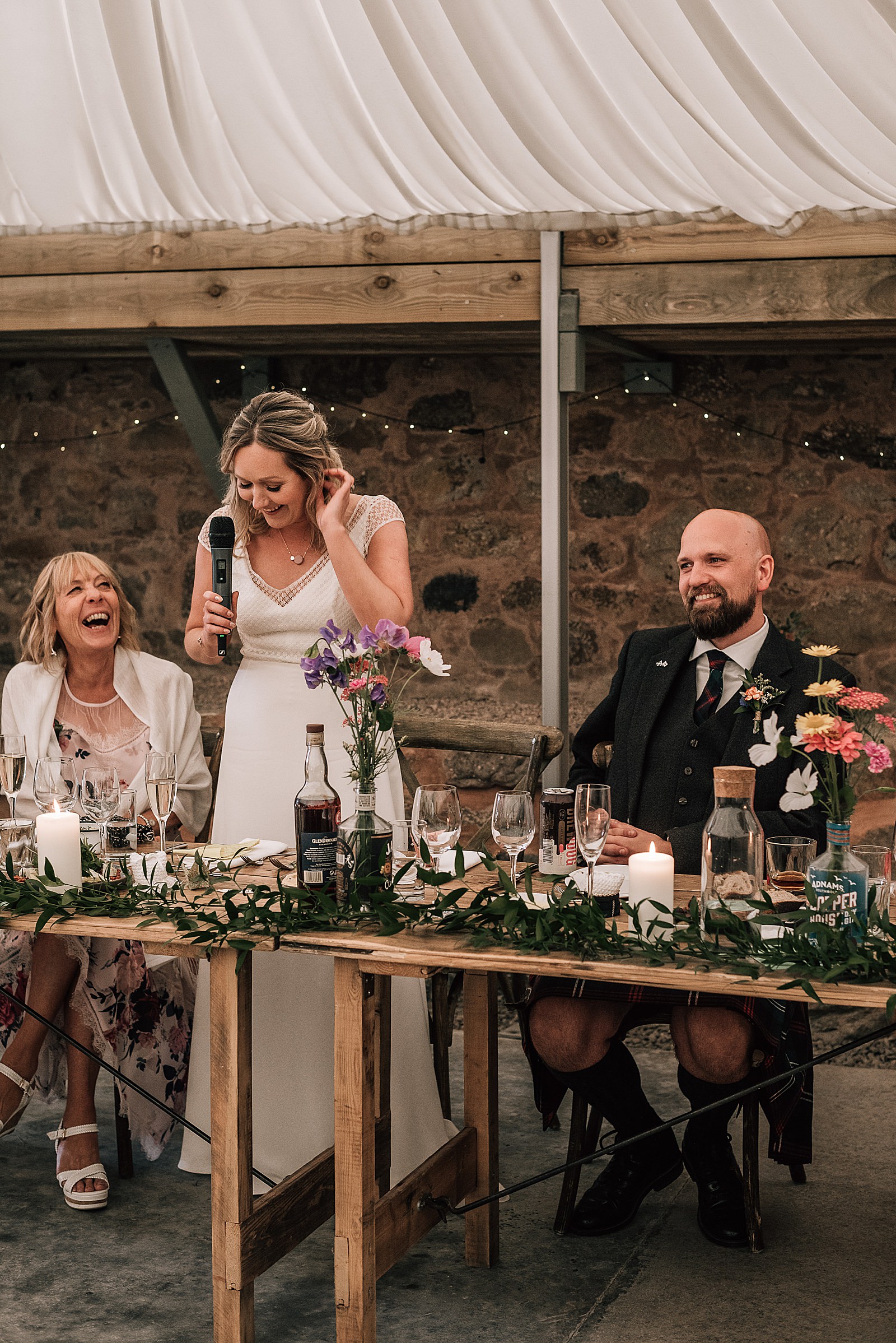 bride giving speech in boho wedding dress at the cow shed scotland with beautiful colourful wild flowers set on a rustic table inside a barn for a diy Scottish wedding