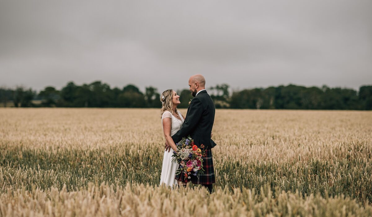 bride and groom standing in wheat field at the cow shed scotland looking at each other smiling after their diy Scottish wedding