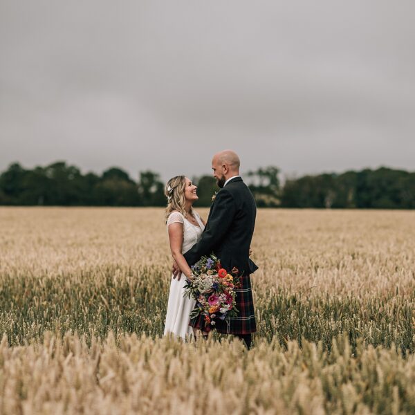 bride and groom standing in wheat field at the cow shed scotland looking at each other smiling after their diy Scottish wedding