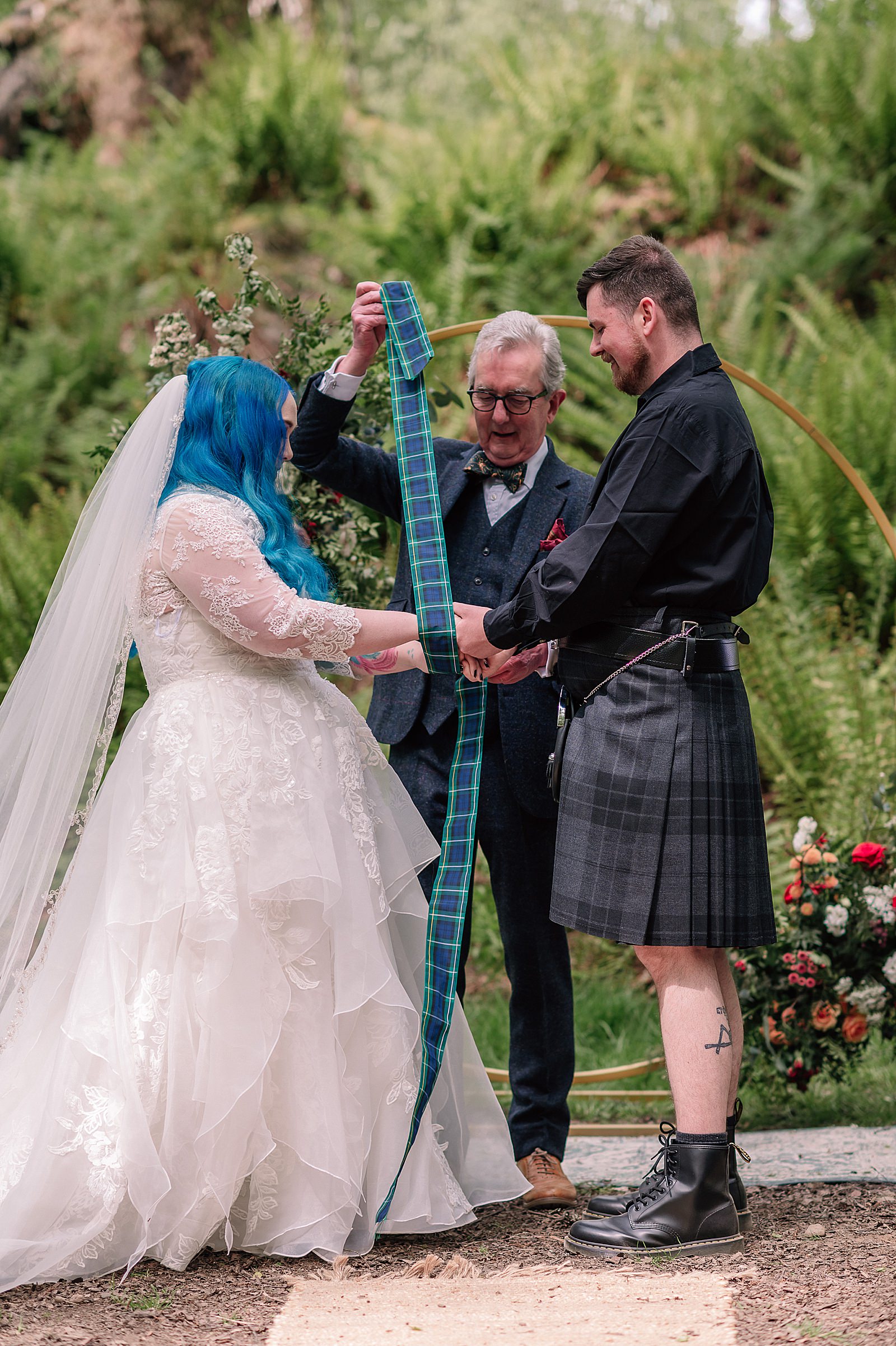 bride with blue hair and groom during hand fasting ceremony with tartan ribbon during their comrie croft wedding ceremony photographed by alternative wedding photographer scotland
