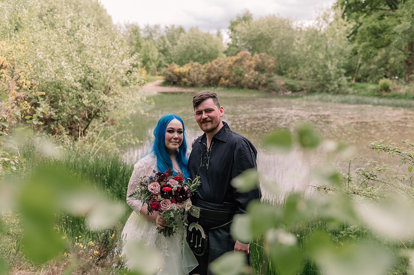 bride with blue hair and groom standing by the side of a pond on a farm for their comrie croft wedding amongst green foliage on a sunny day