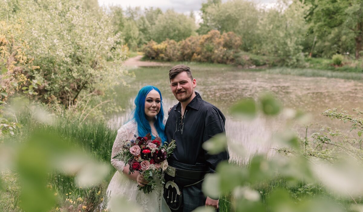 bride and groom standing at the edge of a pond at comrie croft wedding venue surrounded by greenery on a summers day