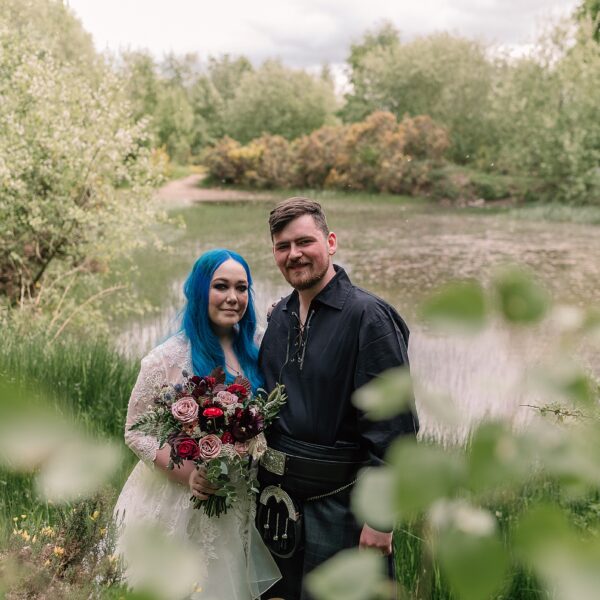 bride and groom standing at the edge of a pond at comrie croft wedding venue surrounded by greenery on a summers day
