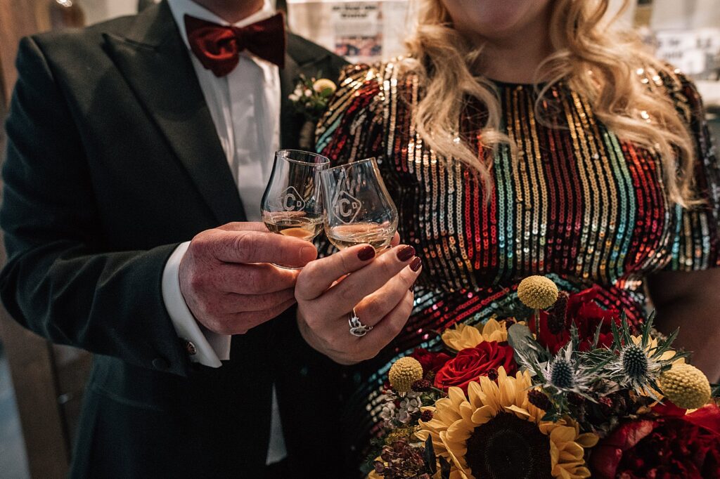 bride in sequin wedding dress with sunflower bouquet and groom cheers with whisky glasses at clydeside distillery for their distillery wedding scotland