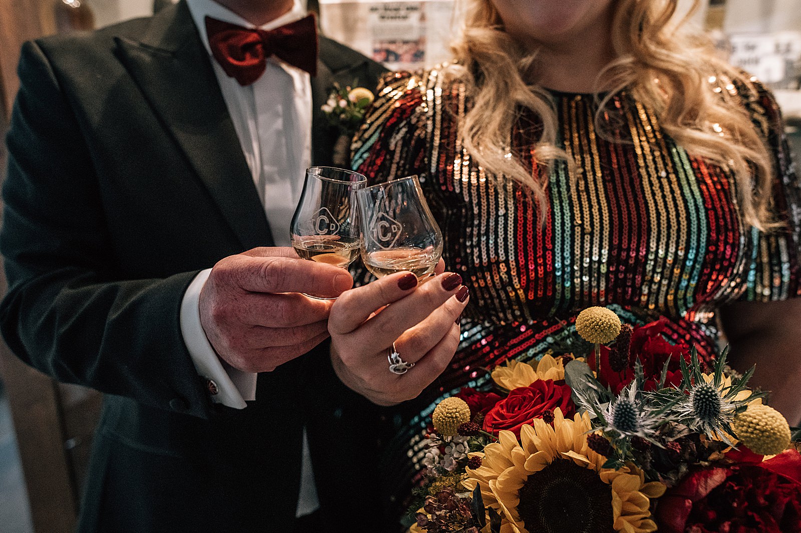 bride in sequin wedding dress with sunflower bouquet and groom cheers with whisky glasses at clydeside distillery for their distillery wedding scotland